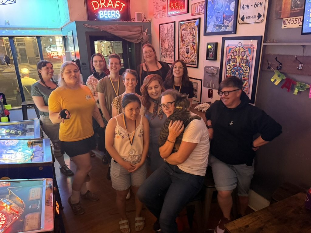 A group of women and a tabby cat gathered in a pinball bar under a neon Draft Beers sign, some wearing medals, one hugging a tabby cat